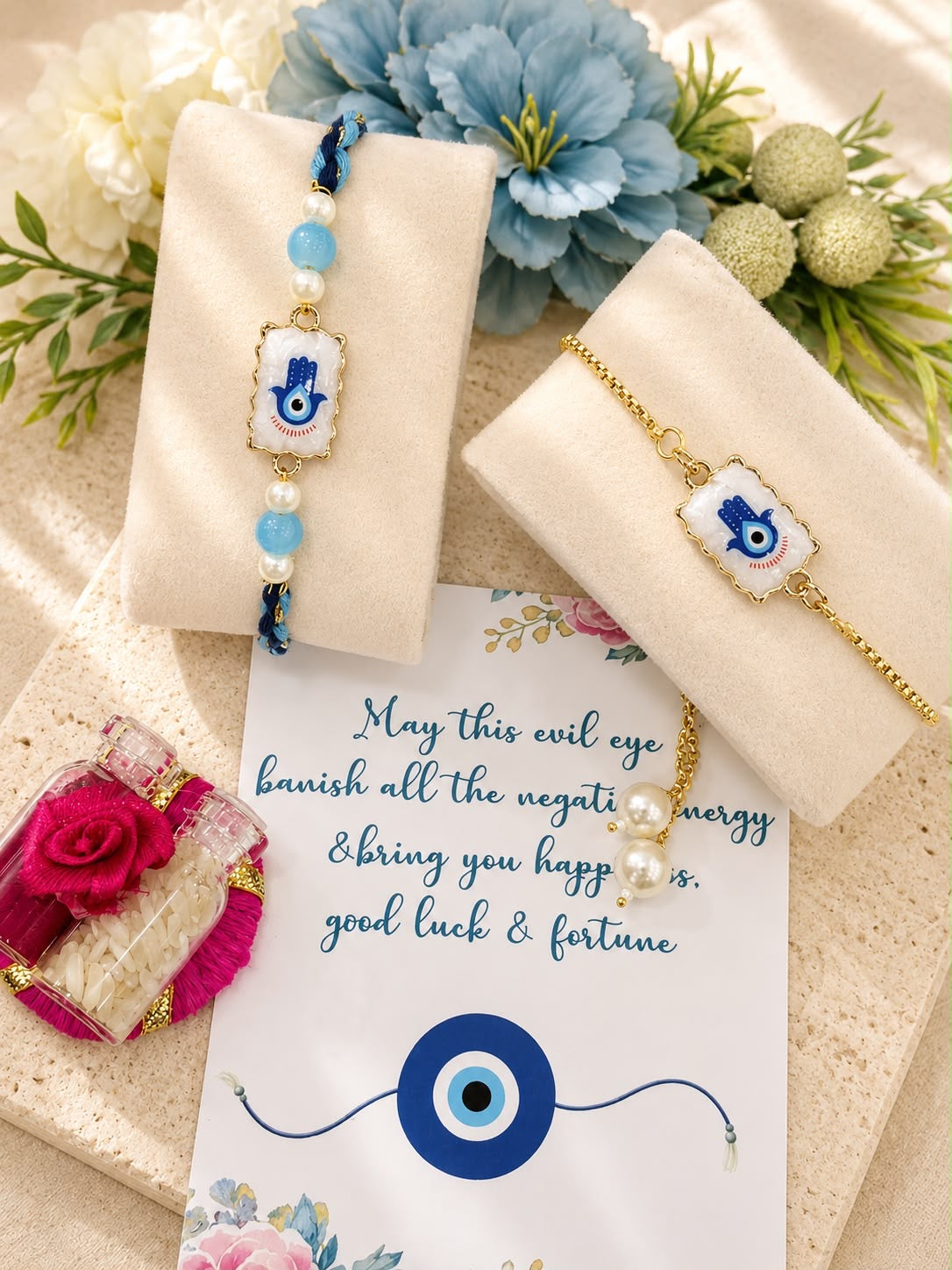 Two evil eye charm bracelets displayed on cream velvet stands, with one blue beaded bracelet and one gold chain bracelet, arranged with flowers and a card featuring a blue evil eye design.