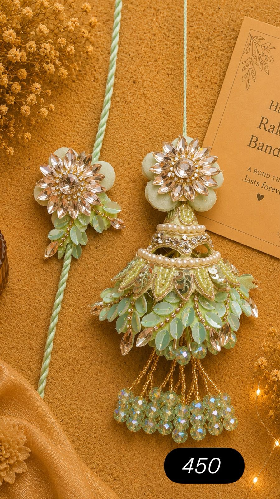 A decorative light green and gold floral garland ornament with rhinestone embellishments, shown alongside a matching floral cord accessory on a warm tan textured background with a card and small lights.