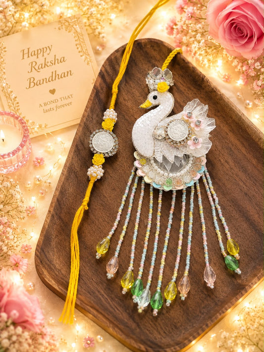 A decorative Rakhi with a white peacock motif, silver embellishments, and dangling multicolored beads, displayed on a wooden tray with a yellow tassel. A matching beaded bracelet and a 'Happy Raksha Bandhan' card are arranged around it with pink roses and warm fairy lights.