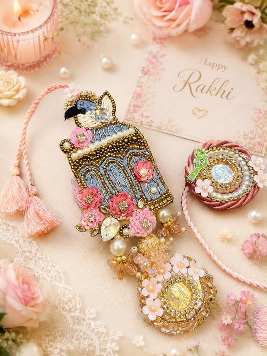 A beautifully arranged flat-lay of traditional Indian Rakhi bangles on a soft beige surface with pink roses, pearls, and a 'Happy Rakhi' card. The central product is a highly detailed, beaded Rakhi featuring a blue and gold bird motif, pink floral embellishments, and a dangling gold-toned ornament with white pearls.