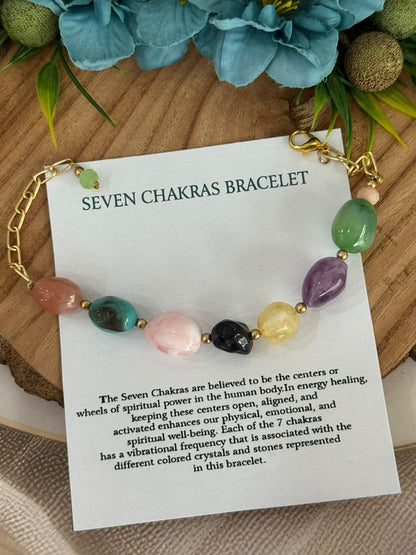 A seven chakras bracelet displayed on a white card with printed descriptions, styled on a wooden surface with blue flowers and green leaves.