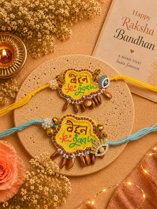 Two decorative Rakhi wristlets displayed on a round beige mat, surrounded by floral props, a lit diya, and a Raksha Bandhan card. One rakhi has yellow thread and gold-tone charms, and the other has light blue thread and gold-tone charms.