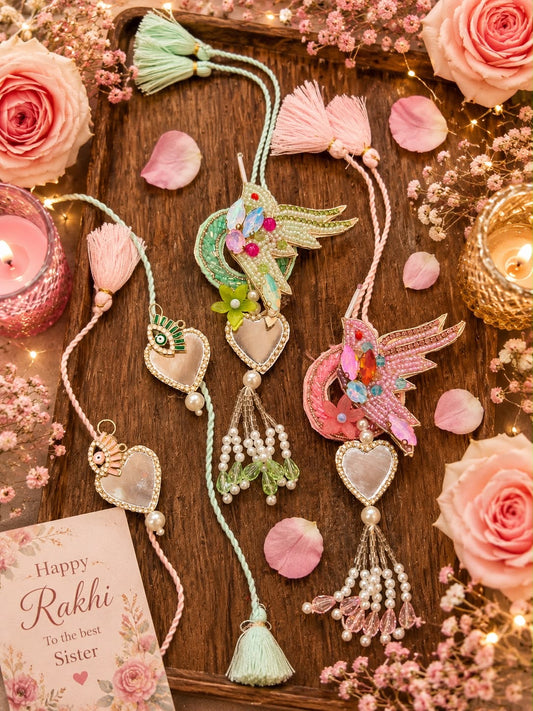 Two decorative Rakhi threads laid out on a wooden tray with pink roses, petals, candles, and a small card reading 'Happy Rakhi to the best Sister.' The product appears to be embroidered Rakhi threads with tassel ends, heart-shaped mother-of-pearl pendants, bead strands, and bird motifs in pastel green, pink, and multicolor accents.