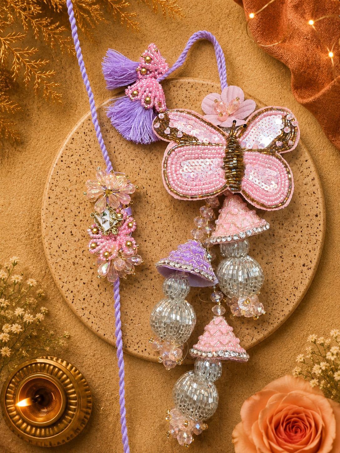 A decorative handmade hanging ornament with a pink sequined butterfly, purple tassels, silver beaded bell-shaped charms, and a lavender cord, styled on a round cork coaster with warm floral and lit-diya props.