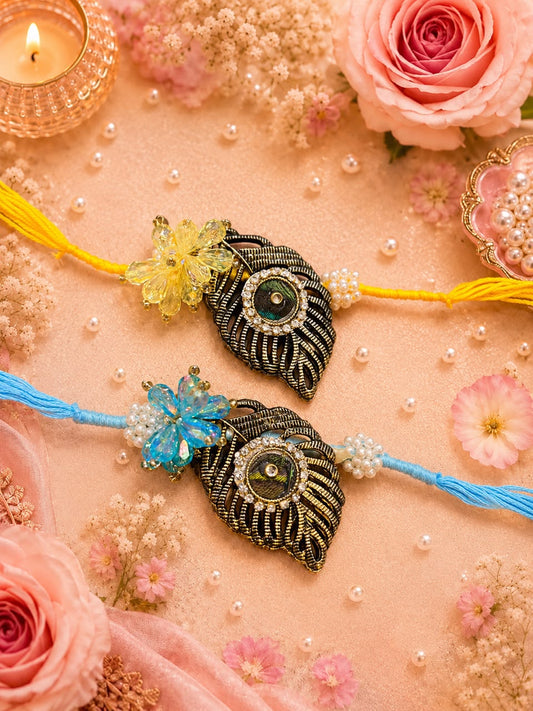 Two decorative Rakhi strands laid on a peach fabric background with flowers, pearls, and a lit candle. The upper Rakhi has yellow thread, a yellow floral embellishment, and a black-and-gold peacock motif with a green stone center.