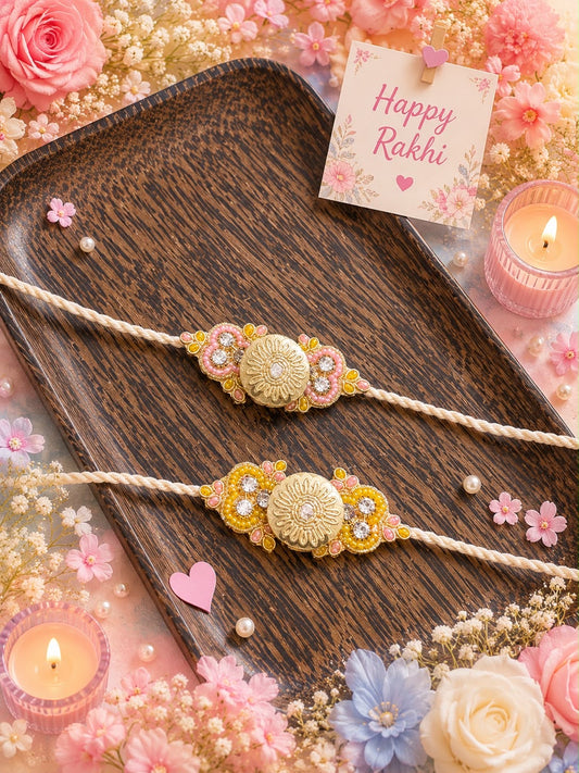 Two decorative Rakhi cords laid on a dark wooden tray, surrounded by pink flowers, candles, and a small card that reads 'Happy Rakhi'. The Rakhis have gold-toned central medallions with pink floral accents and pearl-like embellishments.