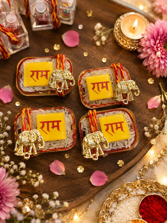 Four rectangular trays of rice decorated with red and orange thread, a small gold elephant figurine, and a yellow square tile printed with the word 'Ram' in red Devanagari script. The trays are arranged on a wooden tray with pink flowers, petals, gold confetti, and a lit candle.
