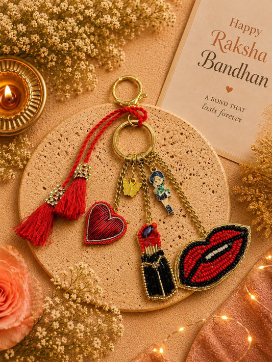 A decorative embroidered keychain or bag charm displayed on a round beige mat, surrounded by dried flowers, a lit diya, and a card reading 'Happy Raksha Bandhan.' The product has red tassels, gold-tone rings and chains, and embroidered charms including a red heart, a lipstick bottle, red lips, and a small blue-and-white figure charm.