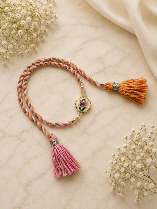 A braided cord bracelet with a central floral charm, featuring one orange tassel and one pink tassel, styled on a cream marble surface with baby's breath flowers.