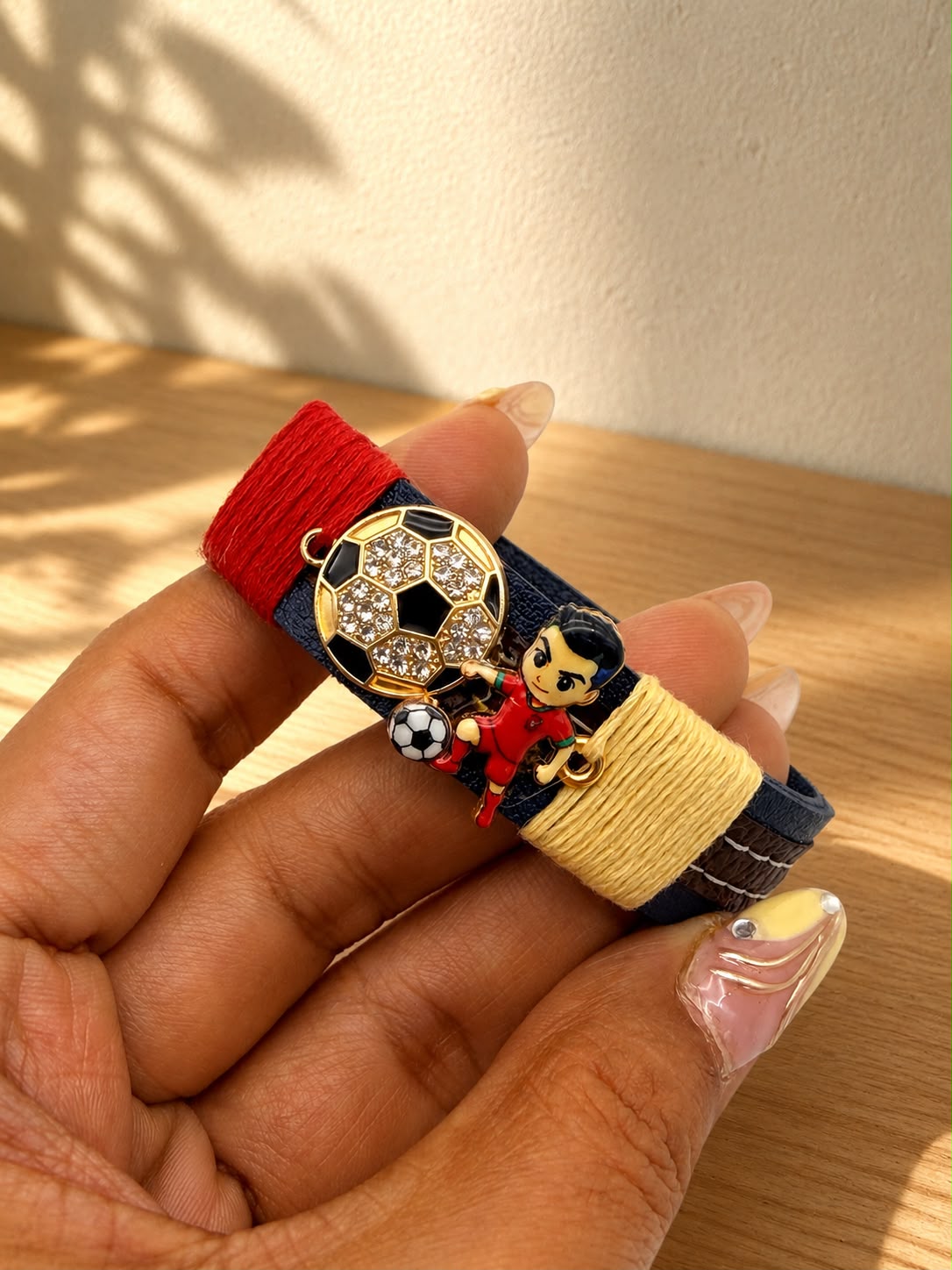 A close-up of a hand holding a colorful bracelet. The bracelet has a navy blue base with a red wrap on one end, a yellow wrap on the other, and a gold-tone soccer ball charm with rhinestones, along with a small character charm.
