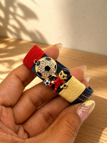 A close-up of a hand holding a colorful bracelet. The bracelet has a navy blue base with a red wrap on one end, a yellow wrap on the other, and a gold-tone soccer ball charm with rhinestones, along with a small character charm.