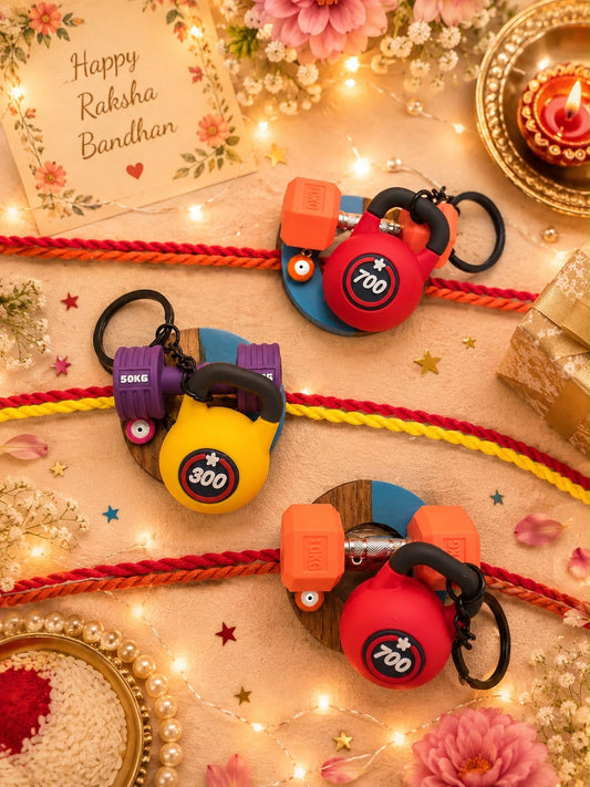 Three colorful Rakhi-themed keychains arranged on a decorated festive surface with red and yellow strings, flowers, lights, and a card reading 'Happy Raksha Bandhan.' The keychains feature small dumbbell-shaped charms in red, yellow, and purple with blue circular bases and black key rings.
