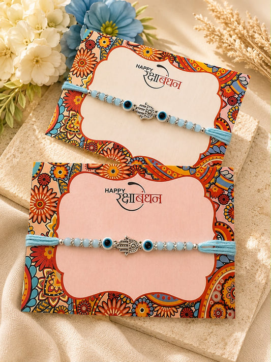 Two traditional Indian Rakhi bracelets with light blue beaded strands and silver-toned central ornaments, displayed on decorative pink and patterned envelopes with flowers and dried wheat in the background.