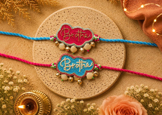 Two handmade Indian Rakhi wristlets displayed on a round cork coaster. The top Rakhi is blue with a magenta cloud-shaped center embroidered with 'Brothers' in gold, and the bottom Rakhi is pink with a turquoise cloud-shaped center embroidered with 'Brothers' in gold.