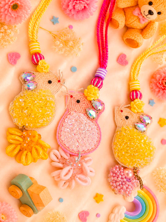 Three handmade owl-shaped keychains or bag charms in yellow and pink, decorated with beads, sequins, and iridescent rhinestones, arranged on a pastel pink background with decorative flowers, a teddy bear, and small toys.