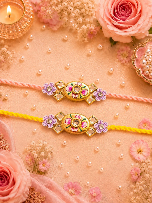 Two traditional Indian Rakhi bracelets laid flat on a peach-colored surface decorated with pink roses, dried flowers, and small pearls. One Rakhi is pink and the other is yellow, both featuring ornate gold-toned floral medallions with pink and purple enamel details.
