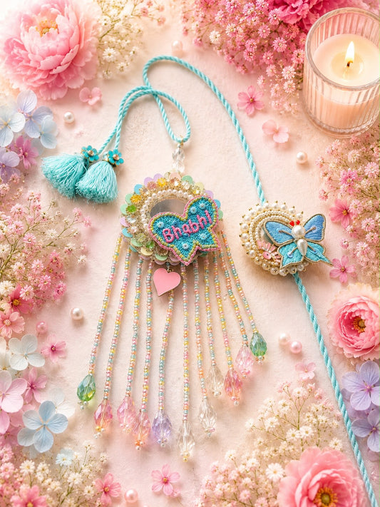 A decorative hanging ornament set styled on a soft pink floral surface. The set includes a turquoise heart-shaped ornament with the word 'Bhakti' in pink, long dangling beaded strands, tassels, and a matching butterfly heart charm on a blue cord.