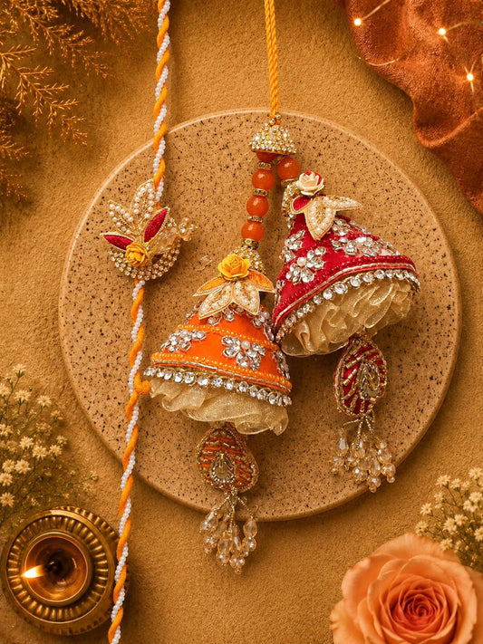 Two ornate decorative hanging ornaments styled like bells, one orange and one red, embellished with silver mirror work, white floral embroidery, and beaded tassels, displayed on a round cork coaster with a braided yellow-orange cord and festive props.