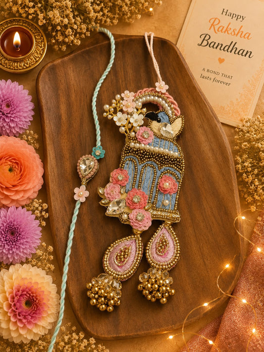 A decorative Rakhi with a matching teardrop pendant is displayed on a wooden board, surrounded by flowers, a lit candle, dried flowers, and a Rakhi Bandhan card.