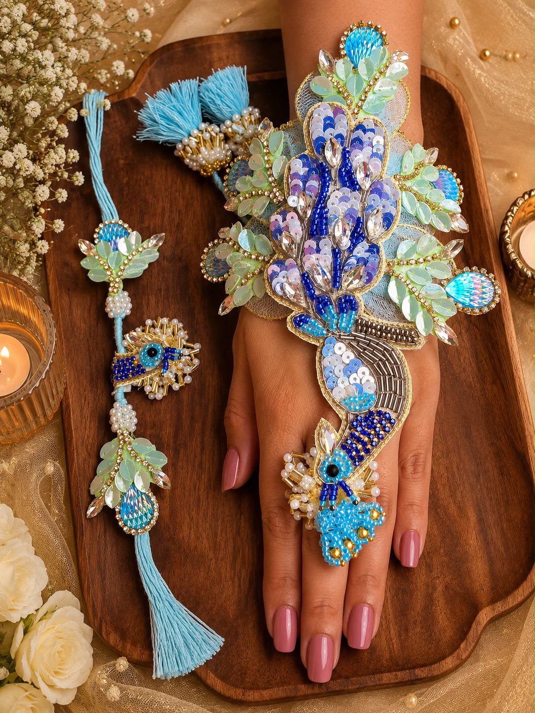 A hand wearing an ornate blue and turquoise peacock-themed jewelry set with matching wrist and toe accessories, displayed on a wooden tray with decorative flowers and candles.