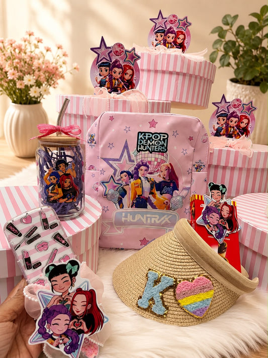 A collection of pink and multicolor K-pop themed stationery and accessories, including a pink striped gift box, a pink zippered pouch with 'K-POP DRAGON HUNTERS' and 'HUNTRX' branding, a clear jar with character stickers, a straw hat with a colorful heart patch, and character stickers, arranged on a white faux fur surface.