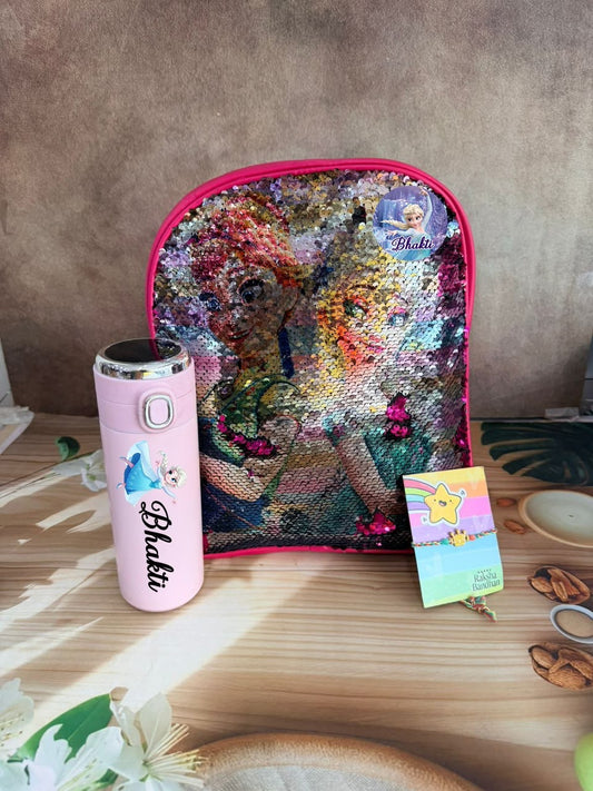 A sequined children's backpack with a colorful reversible design, shown on a wooden table next to a pink insulated bottle and a small card. The backpack features a large sequin pattern depicting a girl with a bow, and a 'Phakti' brand logo is visible on both the bag and the bottle.