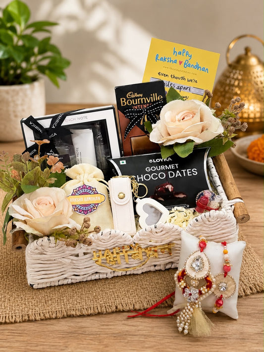 A curated gift basket arrangement on a wooden surface featuring chocolates, flowers, a small pouch, a framed photo, and a Rakhi. Visible packaged products include Cadbury Bournville and Sloya Gourmet Chocolate Dates.
