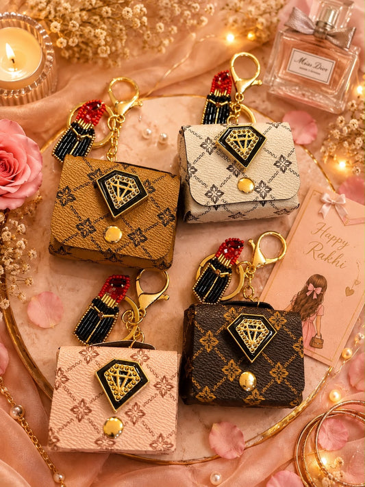 Four mini designer-style keychains or coin purse charms arranged on a pink tray with decorative flowers, candles, and warm lights. The purses have monogram-like patterns in tan, cream, and dark brown, each with a black diamond-shaped front plaque, gold hardware, and a red-and-black tassel charm.
