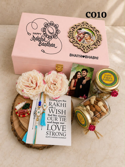 A personalized Rakhi gift set on a light beige fabric background. The set includes a pink wooden box with 'Happy Raksha Bandhan' and 'Bhaviya Bhabhi' printed on it, a photo frame, two glass jars of nuts and sweets, a card with 'Love Strong' text, and two white peony flowers on a wooden slice, all arranged in front of the box.