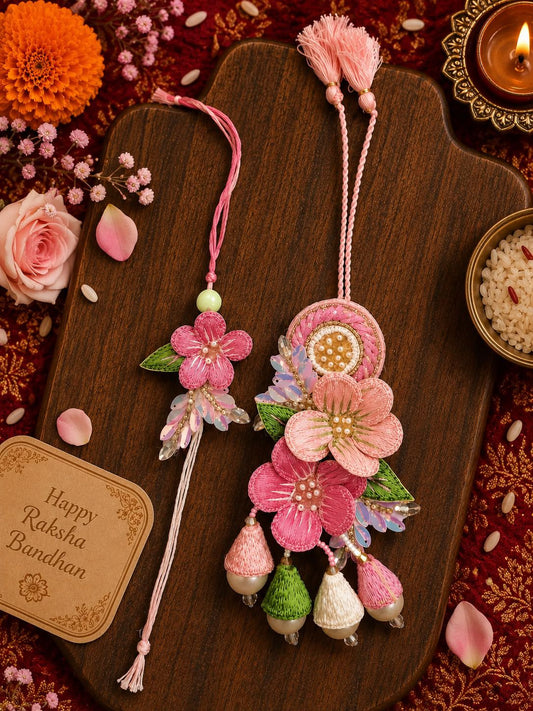 A decorative pink and multicolor floral Rakhi with embroidered flowers, green leaves, and hanging acorn charms, displayed on a dark wooden board with festive props including a lit candle, flowers, and a greeting card.