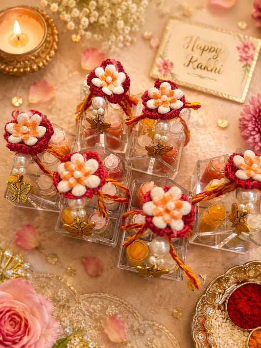 Five small clear gift boxes decorated with red and white flower ornaments, gold foil, and orange tassels, arranged on a beige surface with pink petals, a lit candle, and a card that reads 'Happy Rakhi'.
