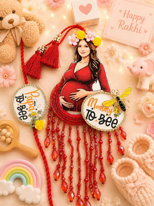 A styled flat-lay of a custom maternity photo plaque featuring a pregnant woman in a red dress with hanging red beads. The plaque is surrounded by baby-themed props including a teddy bear, pink knit booties, a small elephant, a rainbow, and a 'Happy Rakhi' card on a warm beige background.