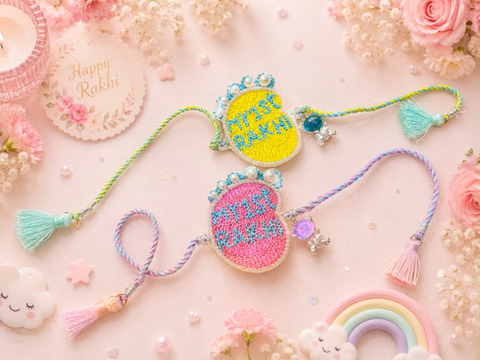 Two decorative embroidered Rakhi bracelets displayed on a pastel pink background with flowers, beads, and a small rainbow charm. One Rakhi is yellow with blue embroidery and the other is pink with blue embroidery, both with colorful cords and tassels.
