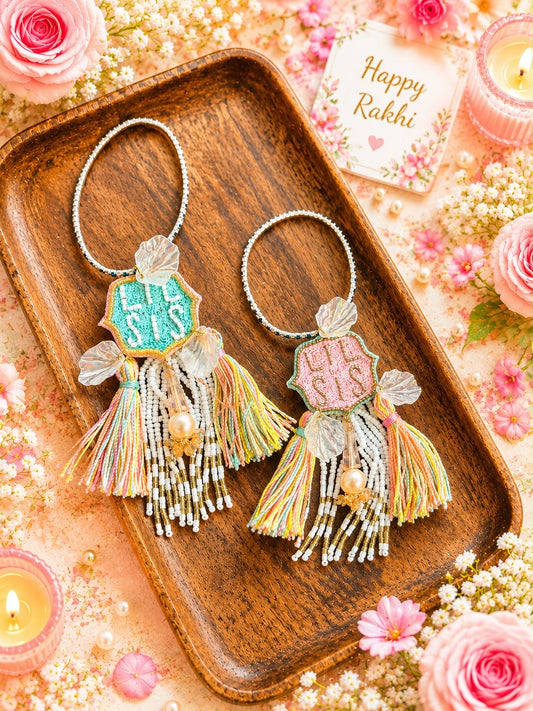 Two handmade Rakhi keychains displayed on a wooden tray, surrounded by pink roses, candles, and a 'Happy Rakhi' card. The keychains feature colorful embroidered 'Rakhi' nameplates, silver beaded rings, and multicolored thread tassels.
