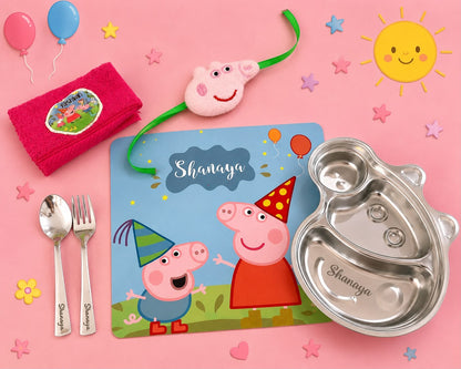 A children's party tableware set on a pink background, including a stainless steel divided plate, a blue printed placemat with Peppa Pig artwork and the name 'Shanaya', a pink plush face mask, a folded pink towel, silver spoon and fork, and balloons.