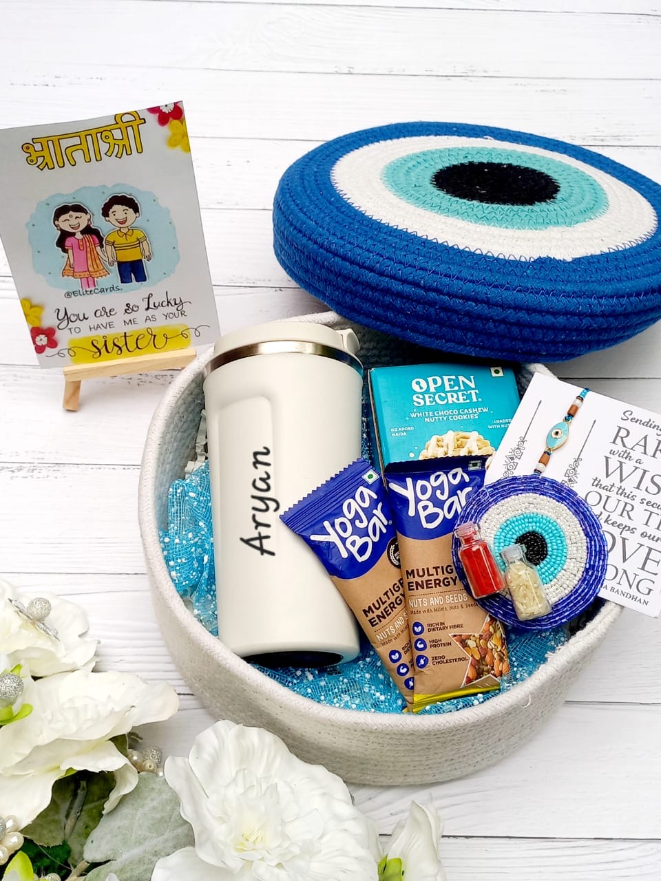 A curated gift basket arranged in a round beige tray with blue glitter filler, featuring a white insulated tumbler with the name 'Aryan', blue and white evil eye coasters, a small card, and packaged snacks including Yogurt Bar and Open Secret. A small printed card on a stand reads 'You are so Lucky.