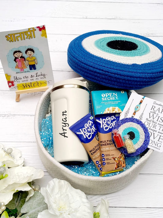 A curated gift basket arranged in a round beige tray with blue glitter filler, featuring a white insulated tumbler with the name 'Aryan', blue and white evil eye coasters, a small card, and packaged snacks including Yogurt Bar and Open Secret. A small printed card on a stand reads 'You are so Lucky.