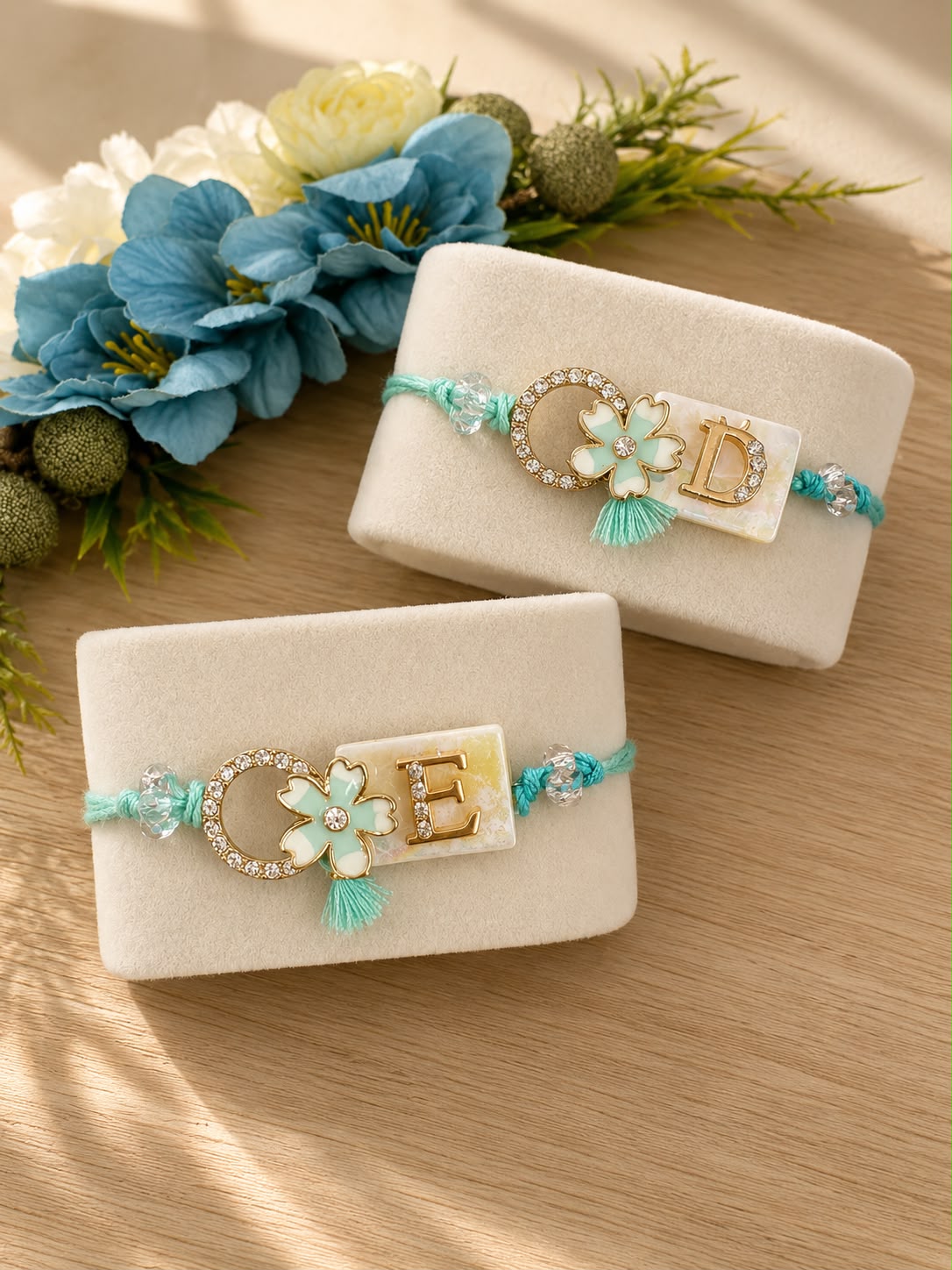 Two personalized braided string bracelets displayed on cream-colored velvet jewelry stands, styled with blue and white flowers on a wooden surface.