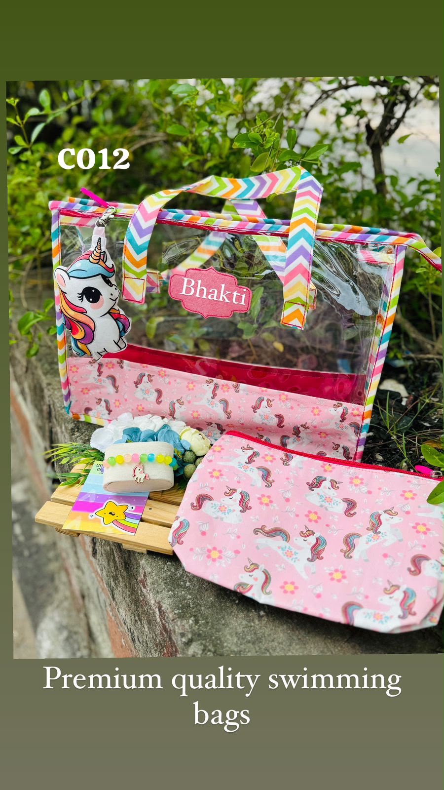 A pink personalized swimming pool bag with a clear front window, rainbow chevron handles, and a unicorn print, displayed outdoors on a stone ledge next to a matching pouch.