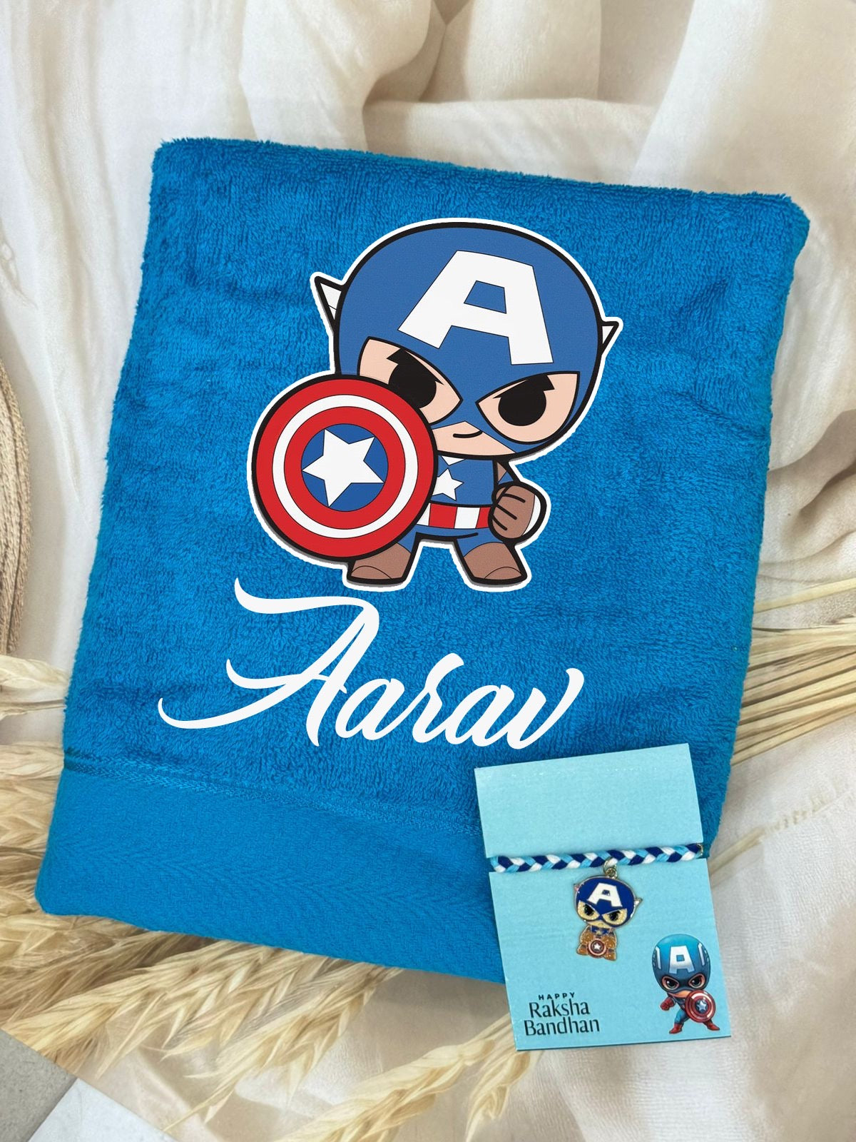 A personalized blue towel featuring a cartoon Captain America graphic and the name 'Aarav' in white script, displayed with a matching card on a beige fabric background.