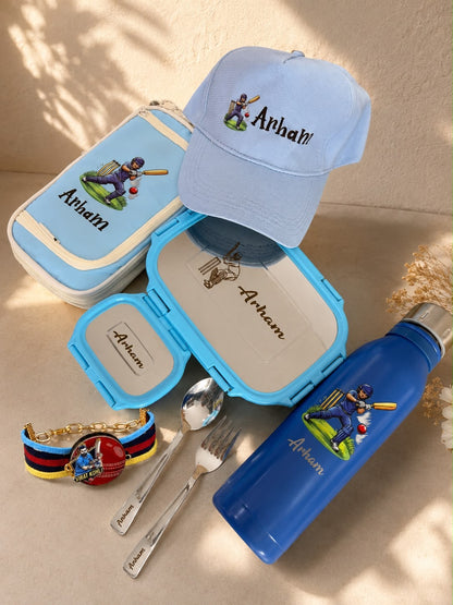 A coordinated set of personalized items featuring the name 'Artem' and a baseball player graphic. The set includes a light blue baseball cap, a light blue lunch box, a medium blue lunch box, a blue water bottle, a blue-and-red beaded bracelet, silver utensils, and a small gold-toned charm, all arranged on a beige surface with soft shadows.