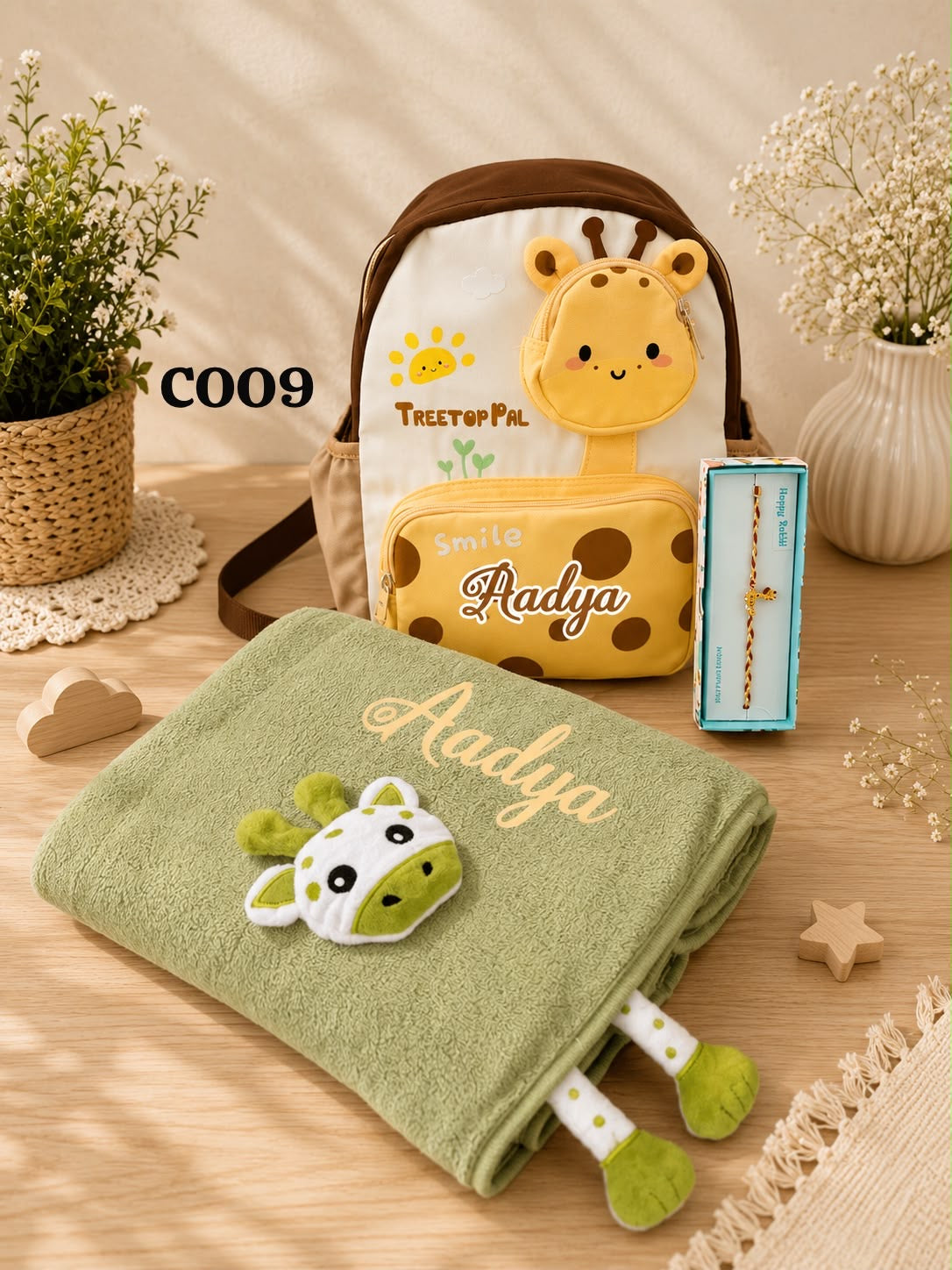 A styled product display featuring a personalized baby towel and matching backpack set. The set includes a sage green folded towel with an embroidered giraffe patch and the name 'Aadya', and a cream backpack with a brown strap and a yellow giraffe face patch, alongside a small yellow zip pouch and a boxed bracelet.