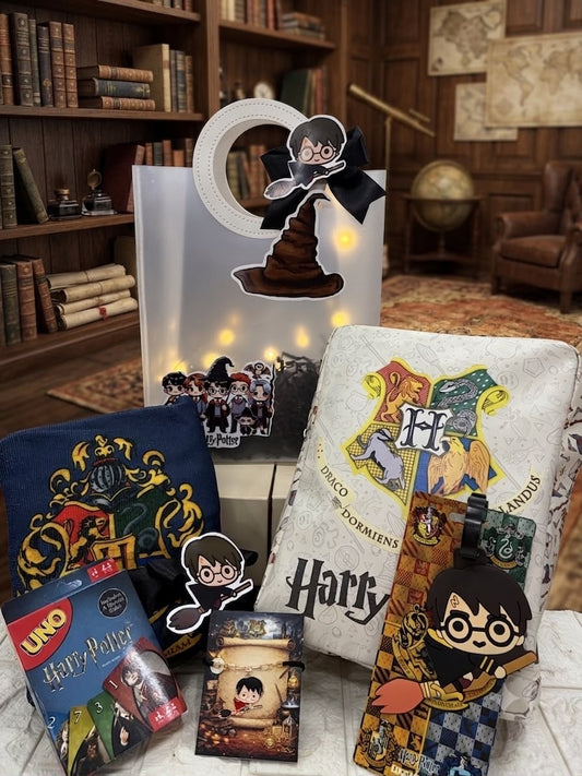 A Harry Potter themed gift set arranged on a tabletop in front of a bookshelf background. The set includes a white Harry Potter gift bag with a Hogwarts crest, a blue Gryffindor-themed pouch, a UNO Harry Potter card deck, a small Harry Potter card or sticker pack, a decorative stand or bag with a Hogwarts silhouette and black ribbon, and several character stickers.