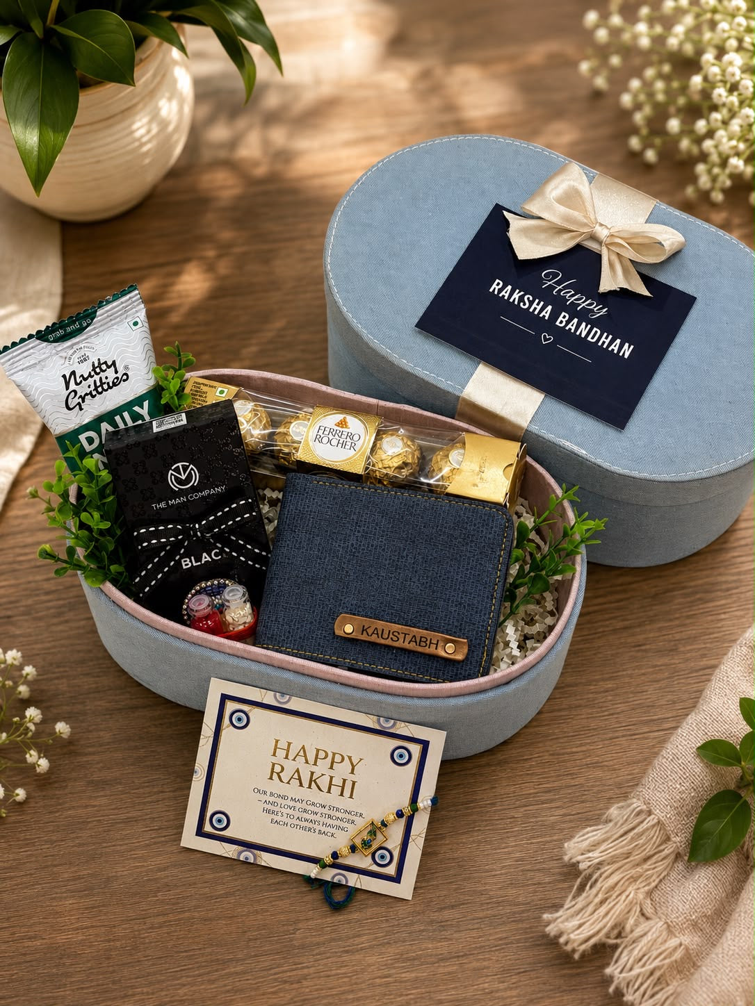A round light blue gift box set on a wooden surface, opened to reveal a wallet, chocolates, and packaged snacks inside. The lid is closed with a cream ribbon and a navy card reading 'Happy Raksha Bandhan.