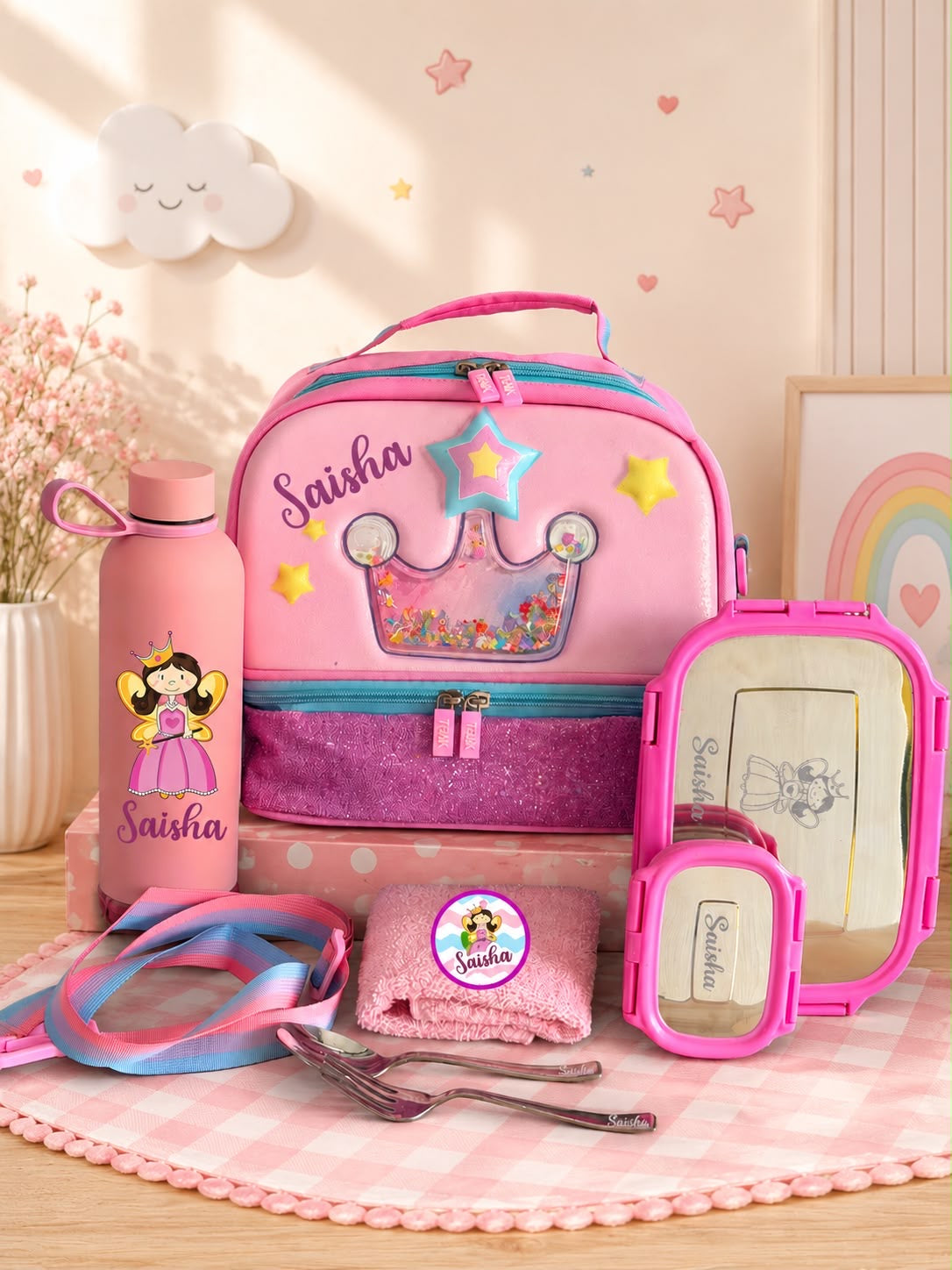 A pink personalized children's lunch set arranged on a pink gingham mat, including a backpack with a crown design, a pink water bottle, a lunch box, a small mirror, a folded towel, and accessories. The items are decorated with the name 'Saisha' and a cartoon princess illustration.