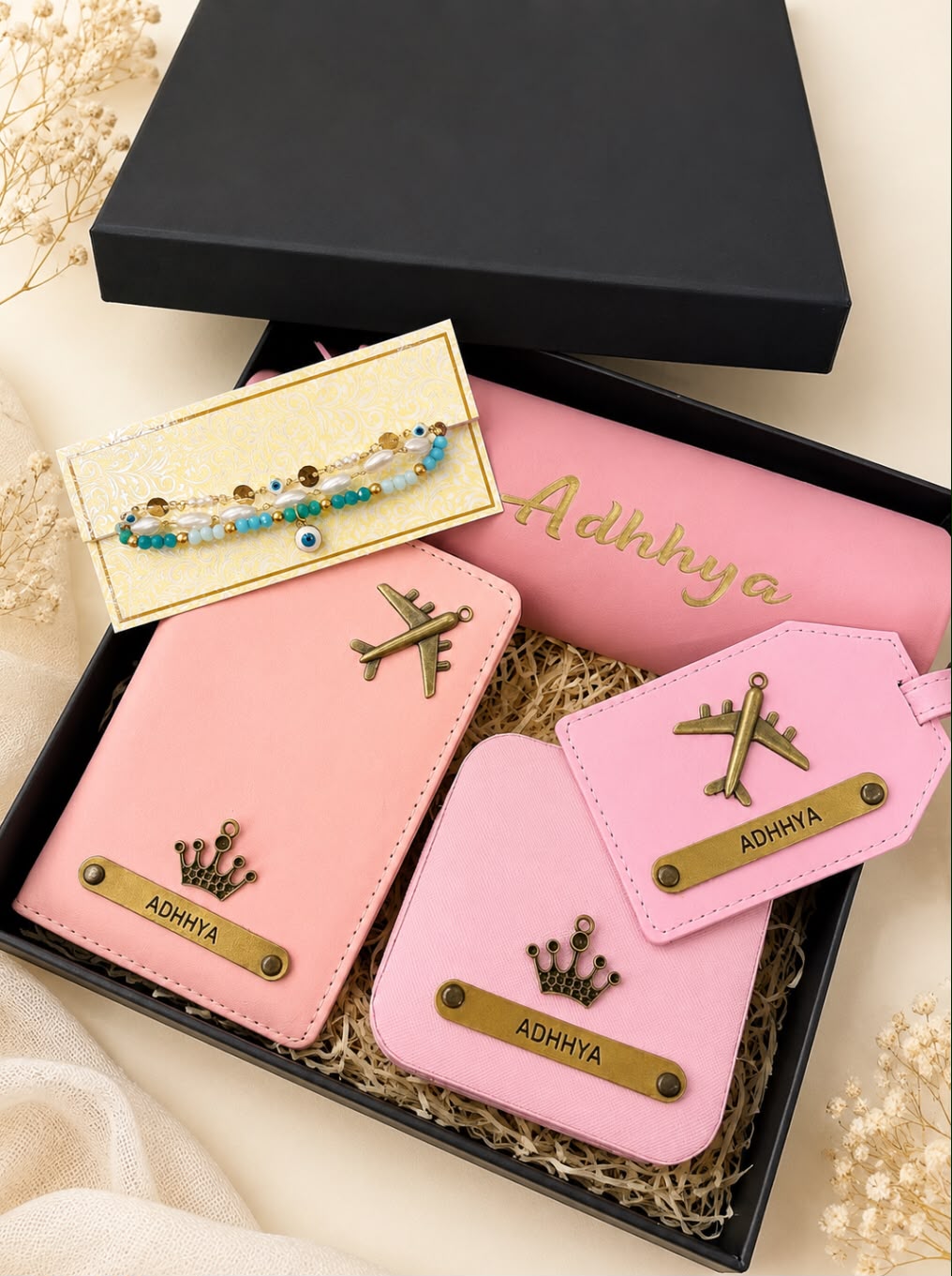 A gift set in a black box featuring pink personalized accessories, including a passport holder, luggage tag, and wallet, with a matching pink pouch, a gold-tone bracelet, and decorative dried flowers on a beige fabric background.