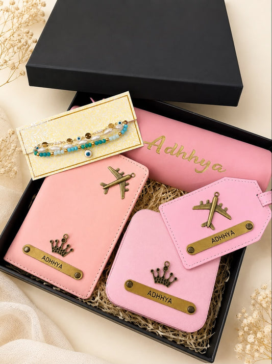 A gift set in a black box featuring pink personalized accessories, including a passport holder, luggage tag, and wallet, with a matching pink pouch, a gold-tone bracelet, and decorative dried flowers on a beige fabric background.
