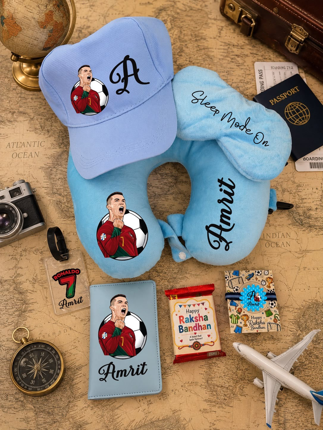 A light blue personalized travel set featuring a baseball cap, U-shaped neck pillow, passport cover, and small card, all embroidered with a soccer player portrait and the name Amrit, arranged on a vintage map background with travel props.