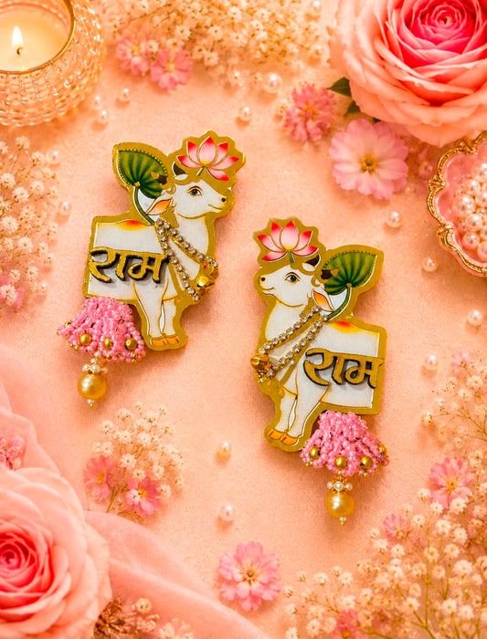 Two decorative Lakshmi enamel pins displayed on a pink fabric background decorated with pink roses, baby's breath, and a lit candle. The pins feature a white cow with pink lotus flowers, gold borders, and the Hindi word 'Ram' written on a pink banner.