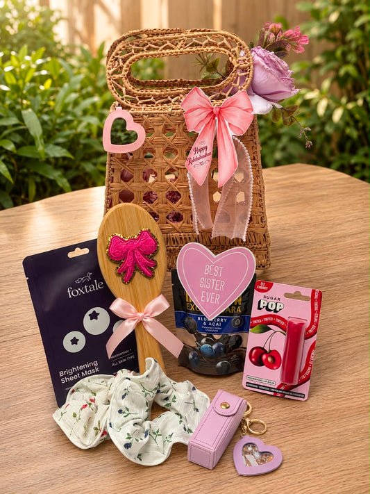 A curated gift basket arrangement on a wooden table featuring a woven tote bag with pink ribbon and heart charms, a wooden spoon with a pink bow, a pink heart-shaped card, a small pink box, a floral scrunchie, and packaged accessories, with a blurred garden background.